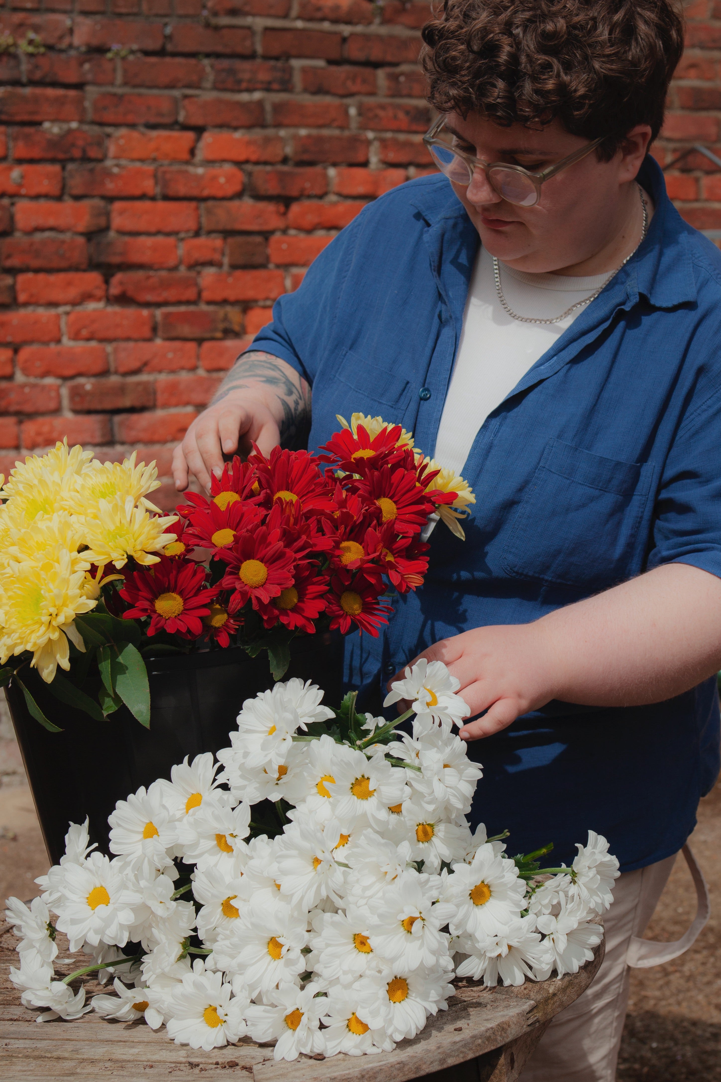A person in a blue work shirt and glasses is arranging a large black planter filled with yellow, red, and white daisy-like flowers on a rustic wooden table in an outdoor garden setting with a brick wall background.